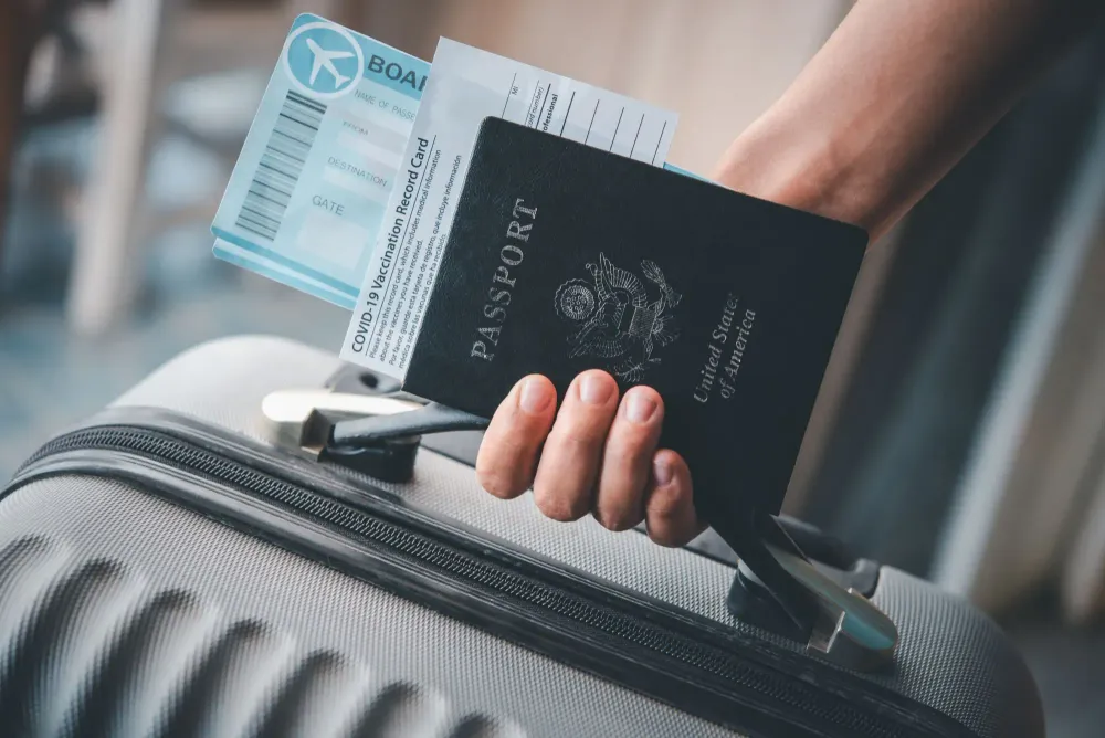 A person holding their COVID-19 vaccine card, passport, boarding pass, and suitcase